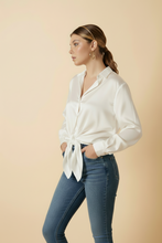 Load image into Gallery viewer, Woman wearing a white blouse with a tie detail and blue jeans on a beige background